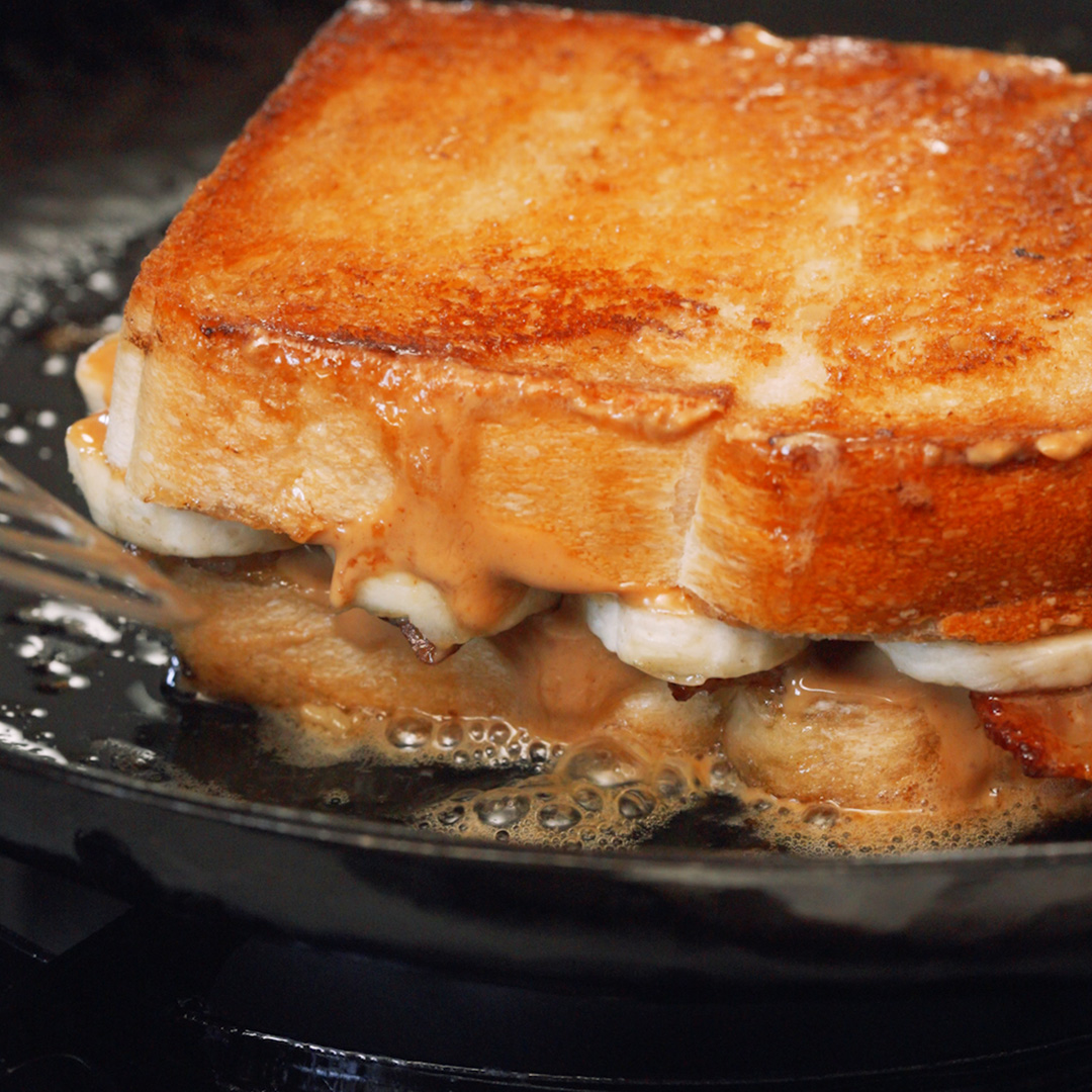 a peanut butter, banana and bacon sandwich being fried in butter on a carbon steel skillet