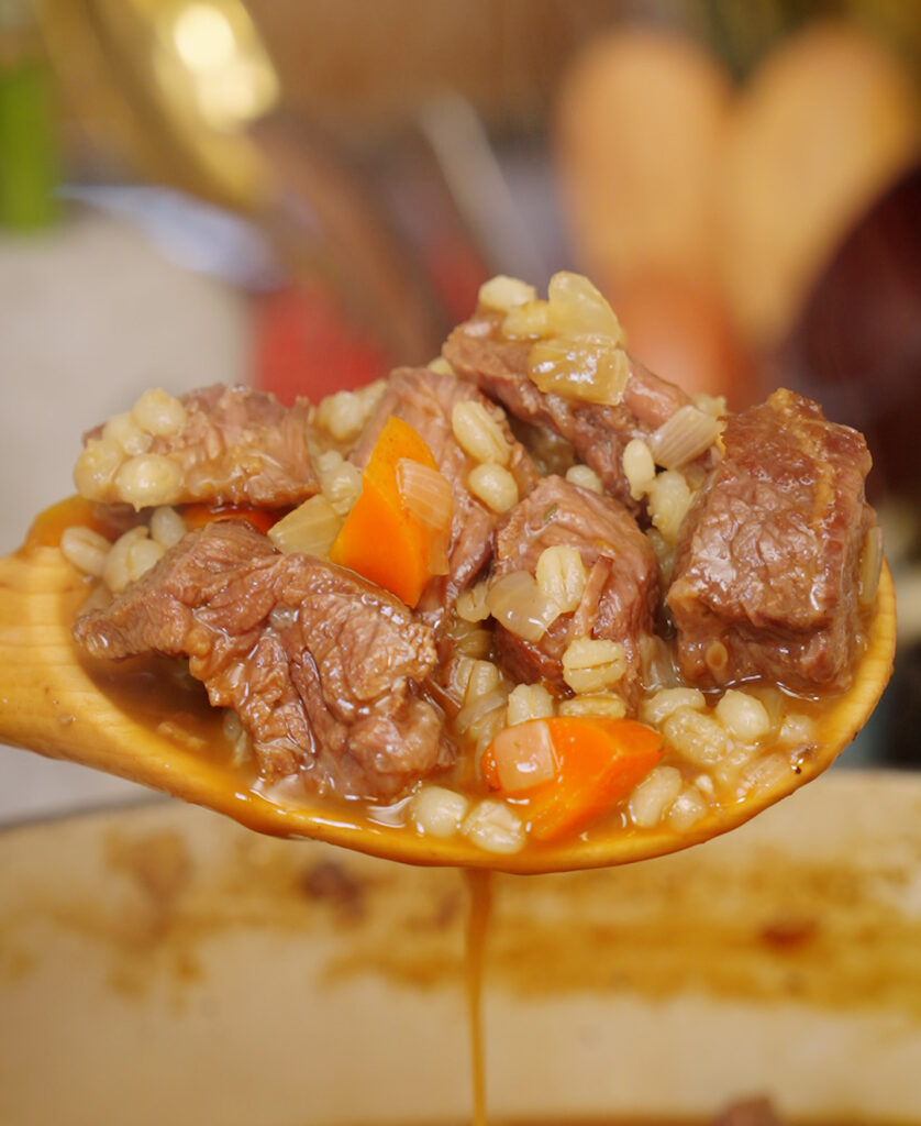 a ladle full of beef barley soup featuring beef chunks, barley and chopped carrots in broth.