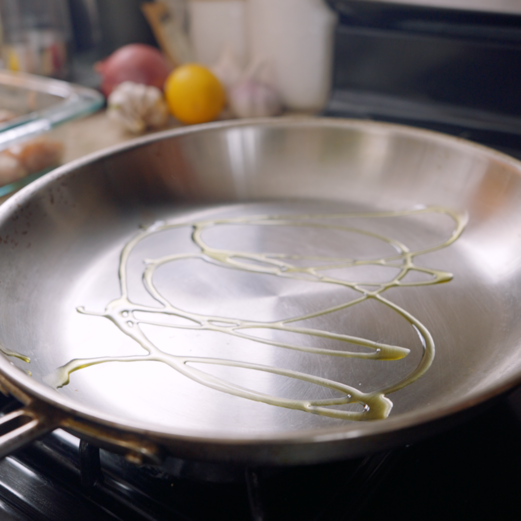 Oil is drizzled across the surface of an empty stainless steel skillet set on a stovetop.