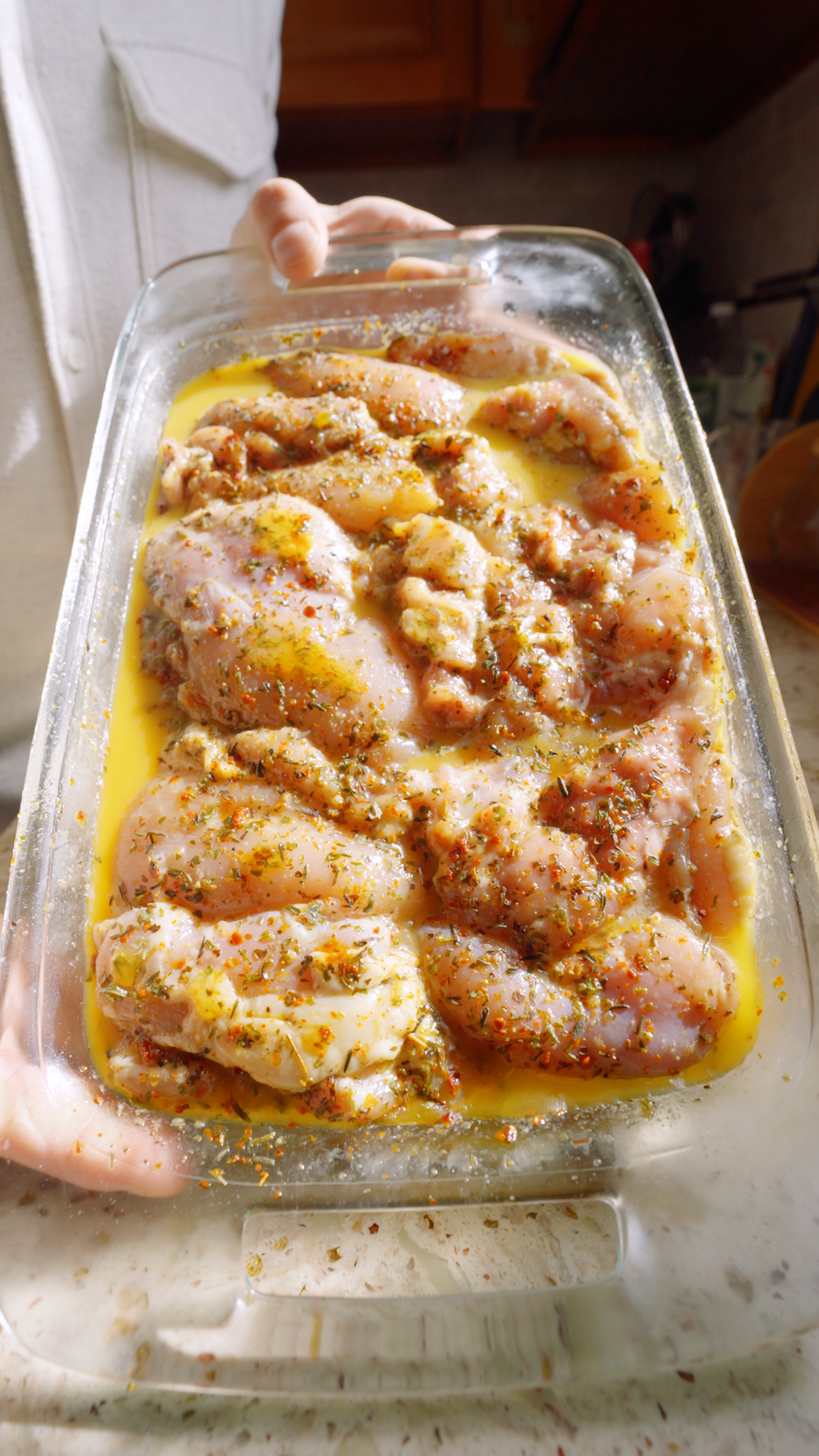 Raw chicken thighs sit in a glass baking dish filled with a yellow marinade, with dried herbs and red pepper flakes visible on the meat and in the liquid.