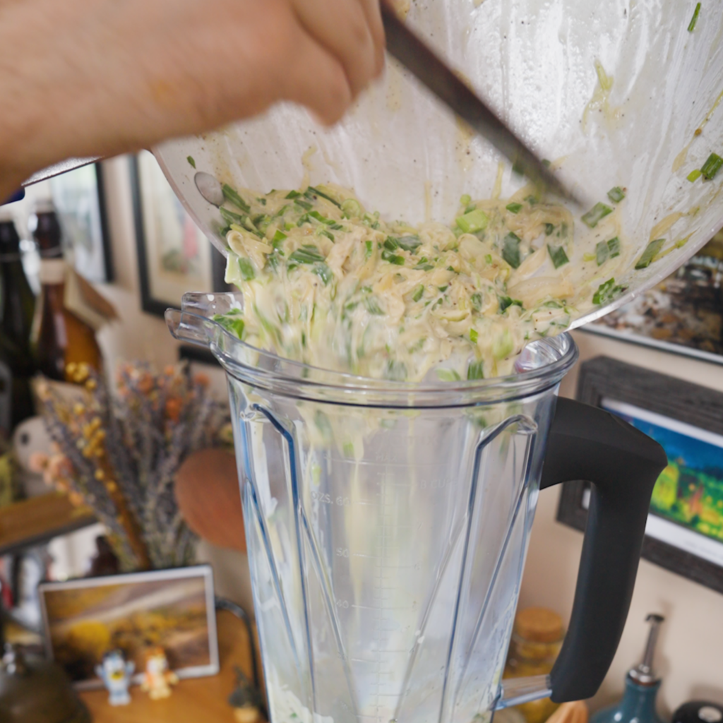 A creamy spring onion mixture with visible sliced onions and green pieces is poured from a pan into a blender.