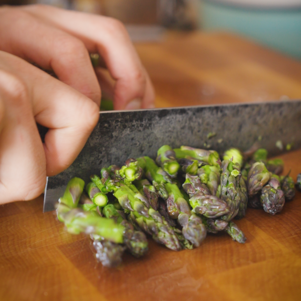 A knife chops fresh asparagus into small pieces on a wooden cutting board, showing green stalks and purple-tipped ends.