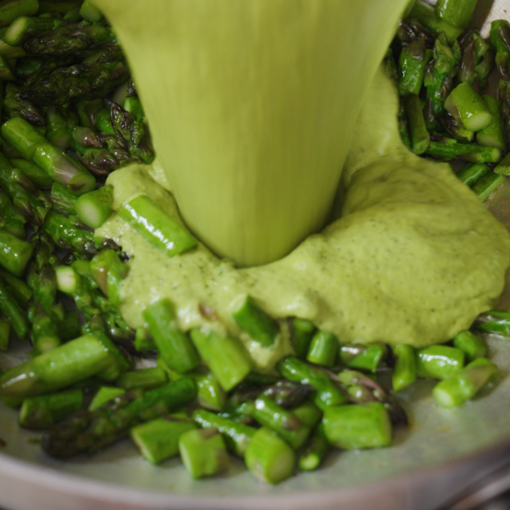 A bright green creamy sauce is poured into a pan of sautéed asparagus pieces, pooling and spreading around the vegetables.