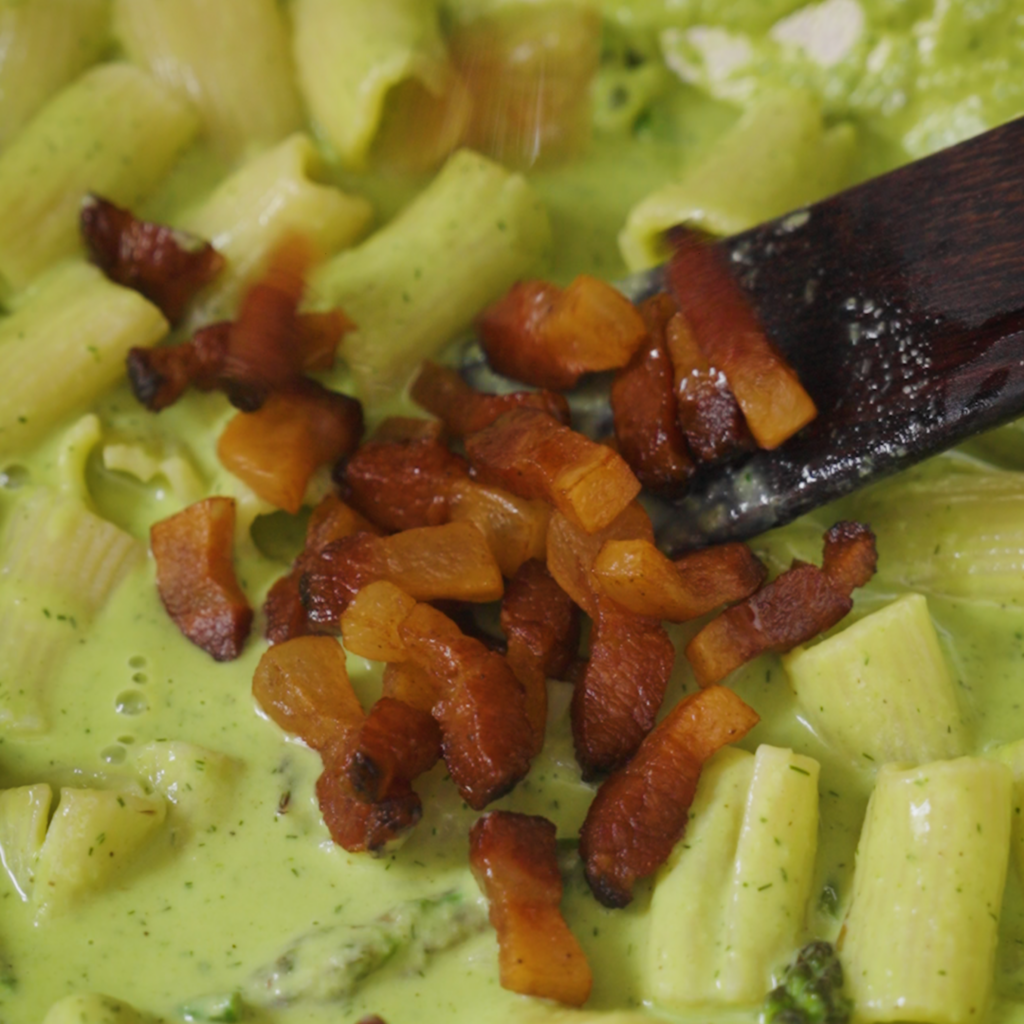Crispy browned guanciale pieces are added to a pan of rigatoni coated in a creamy green sauce.