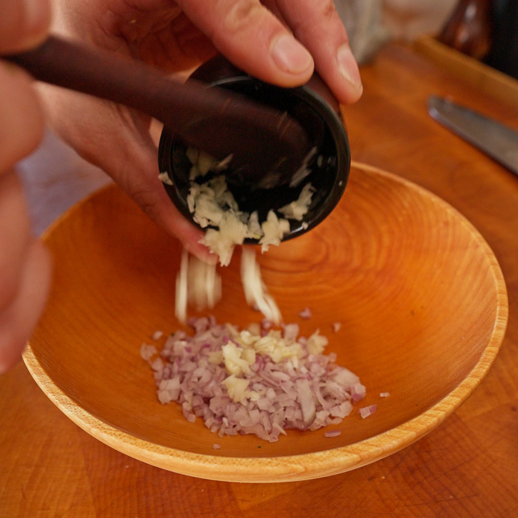 Minced garlic being scraped from a small black bowl into a wooden bowl containing finely diced shallot on a wooden countertop.