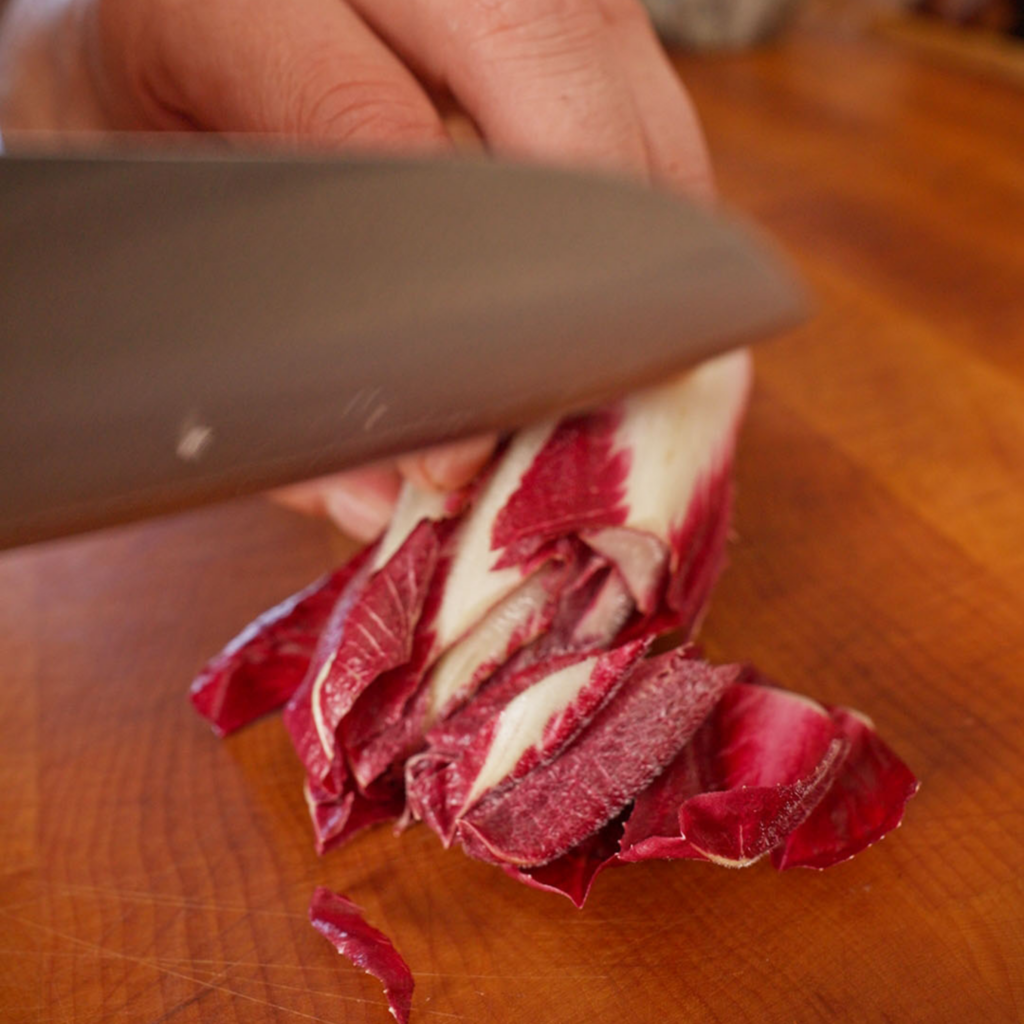 A chef’s knife slicing a head of red endive into thin strips on a wooden cutting board.