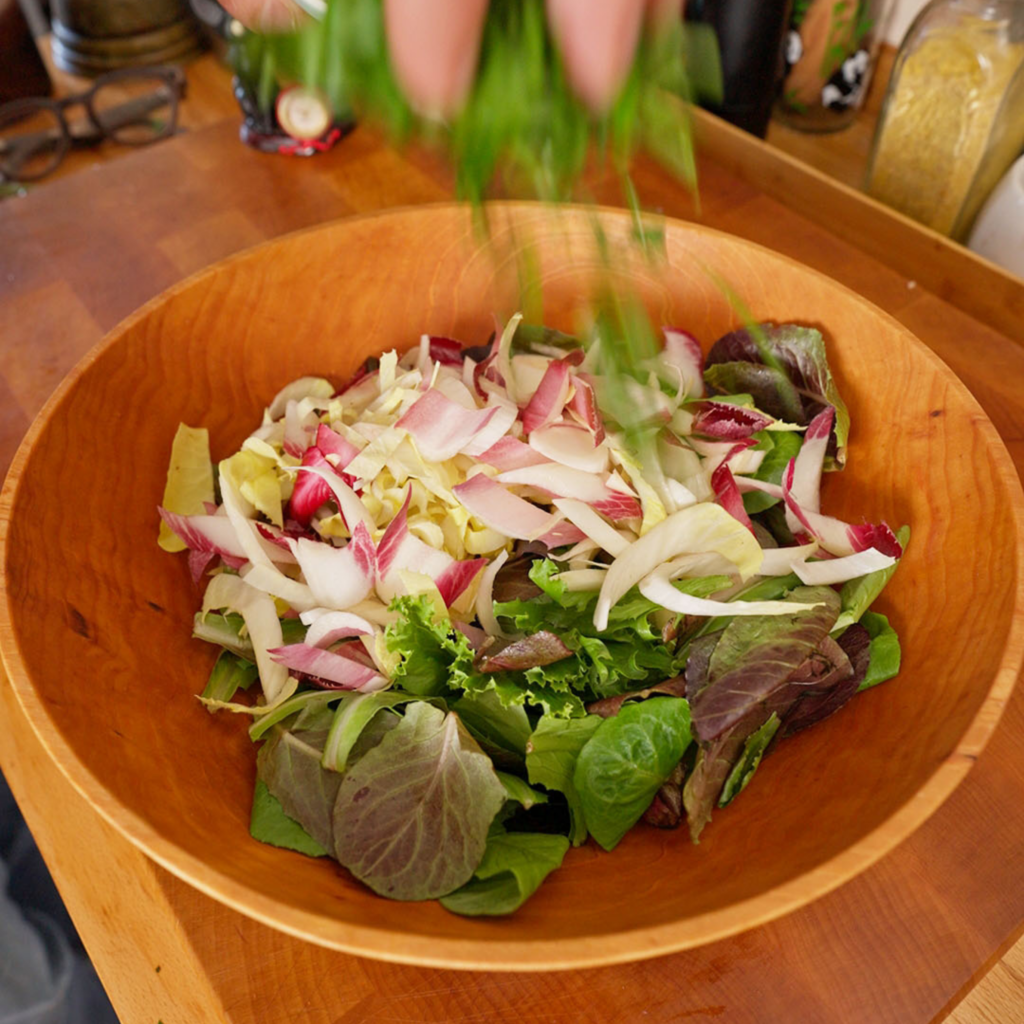 Mixed salad greens and thinly sliced endive in a wooden bowl as a hand drops in fresh herbs from above.