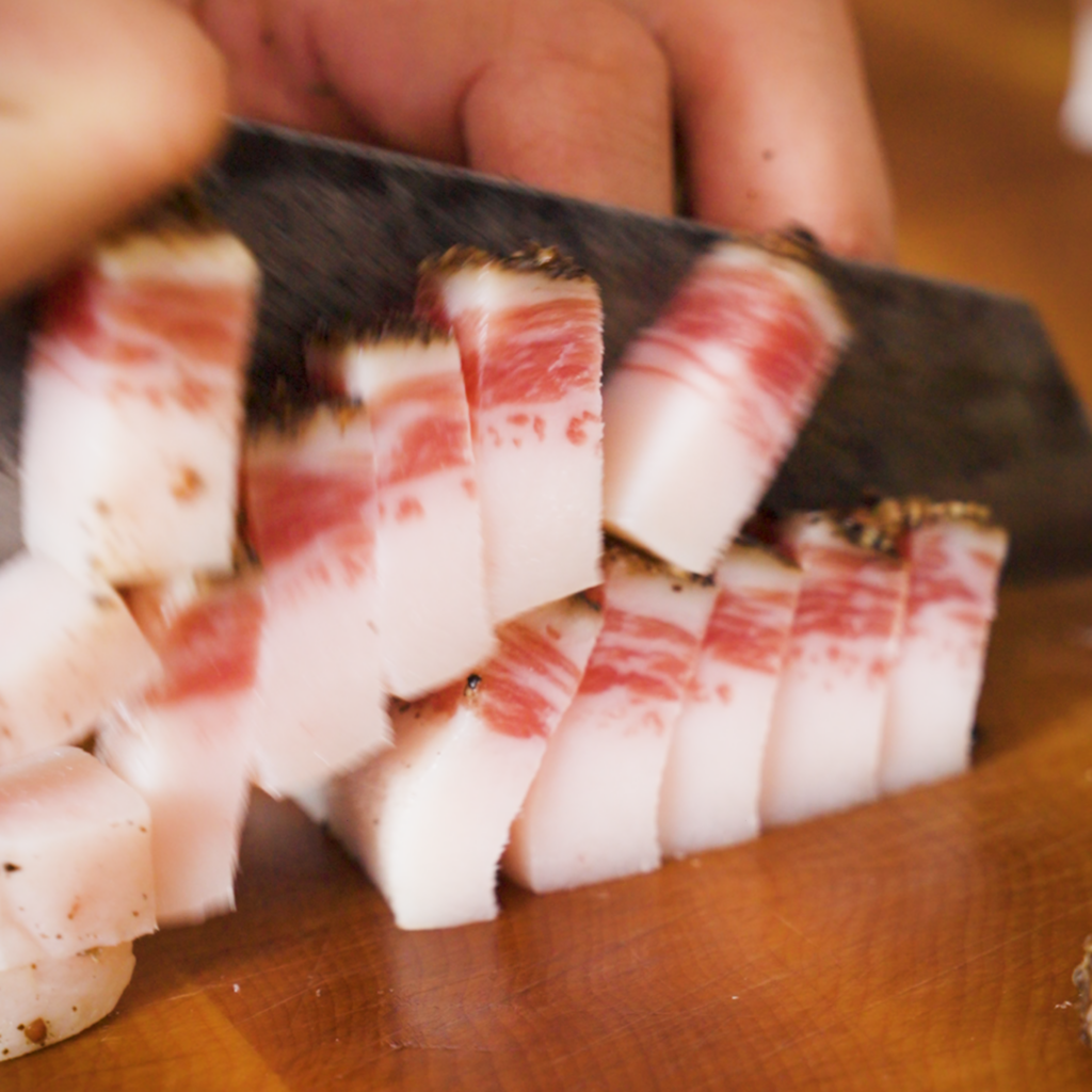 Close-up of thick-cut guanciale being sliced into lardons on a wooden cutting board, showing distinct layers of white fat and pink meat.