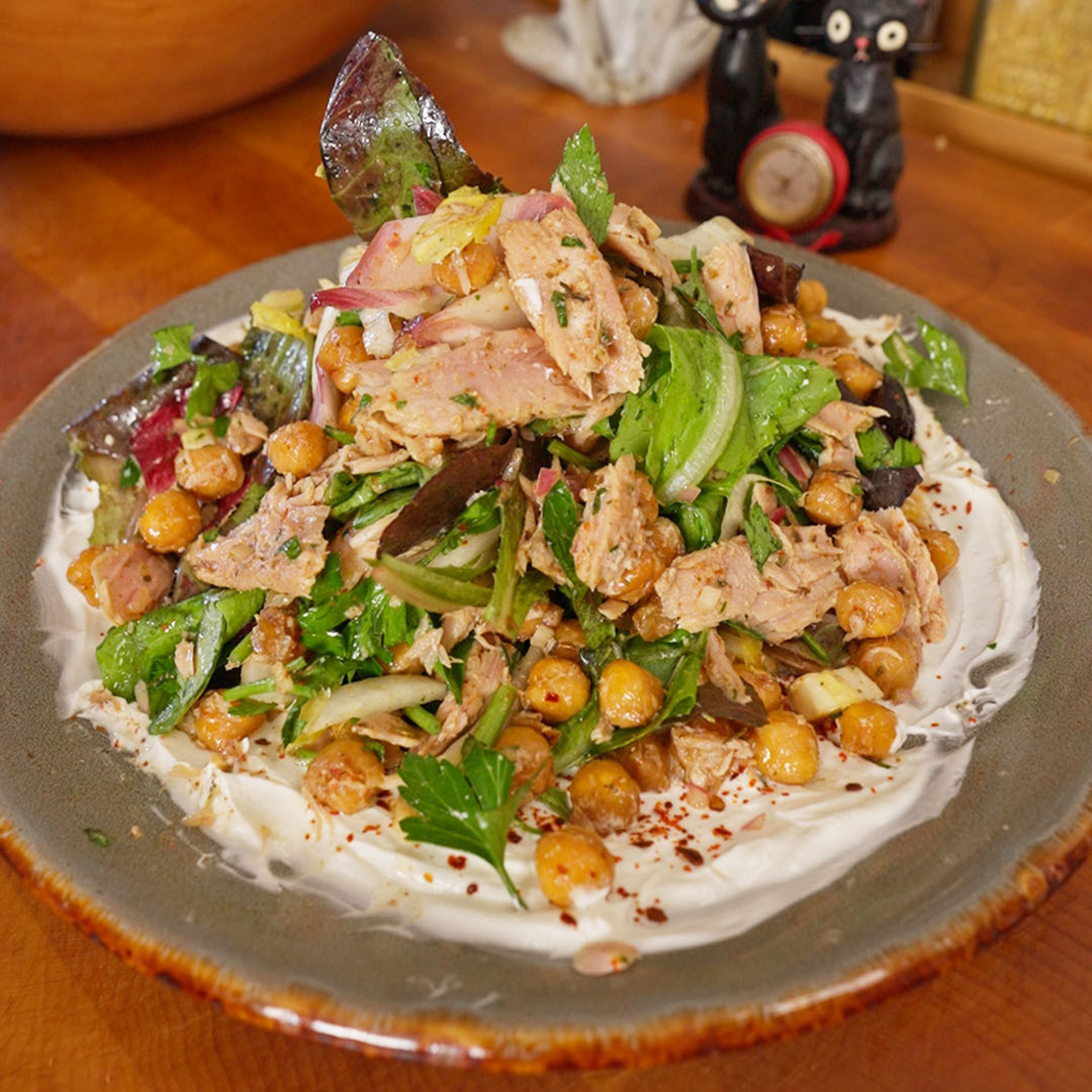 A ceramic plate with a thick layer of yogurt topped with flaked tuna, crispy chickpeas, mixed greens, sliced red onion, and chopped herbs, with red pepper flakes scattered on top.
