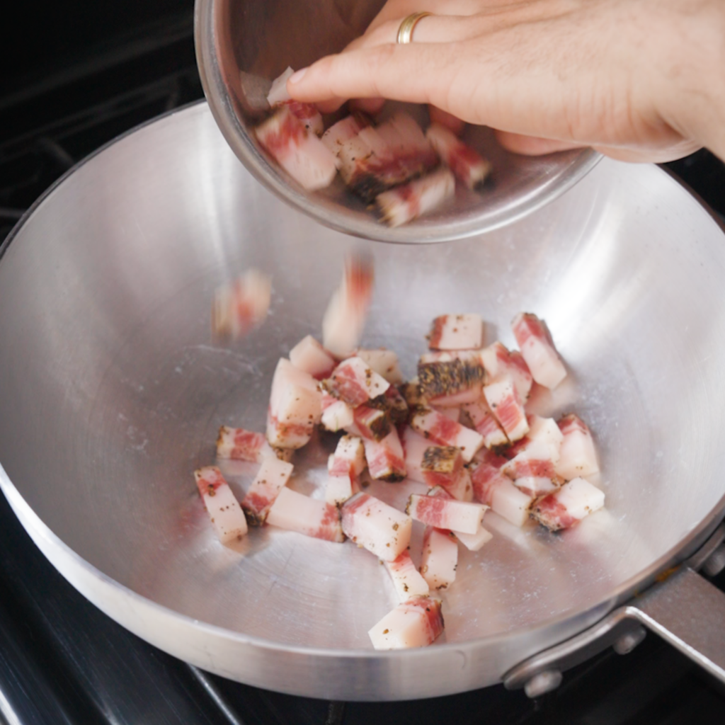 A hand pours guanciale from a metal bowl into a large stainless steel pan on a stovetop.