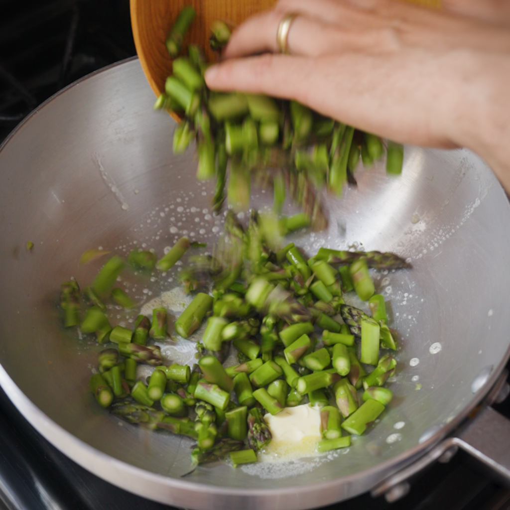 Chopped asparagus is added to a stainless steel pan with melting butter on a stovetop.