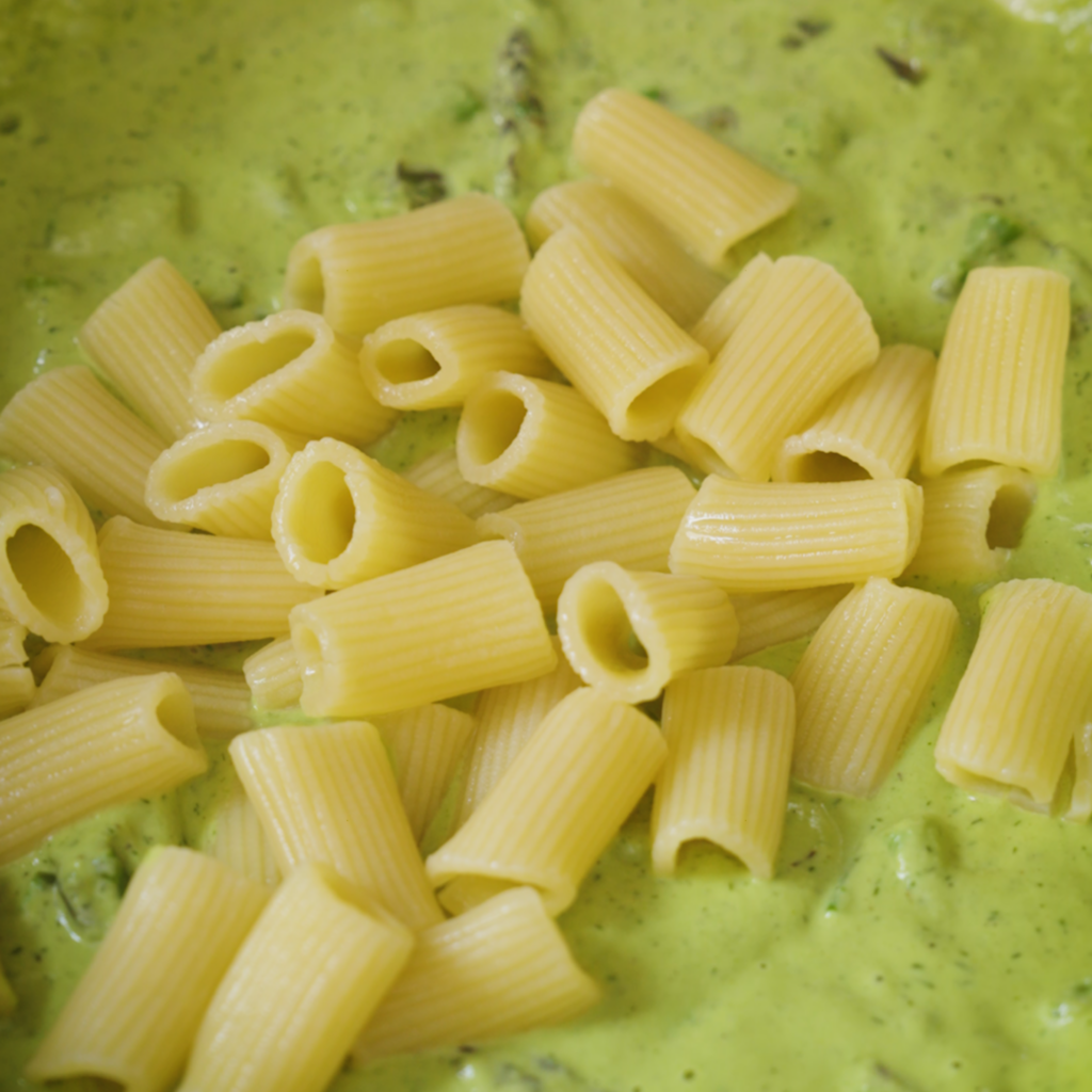 Cooked rigatoni pasta is added to a pan filled with a smooth green sauce with visible asparagus pieces.