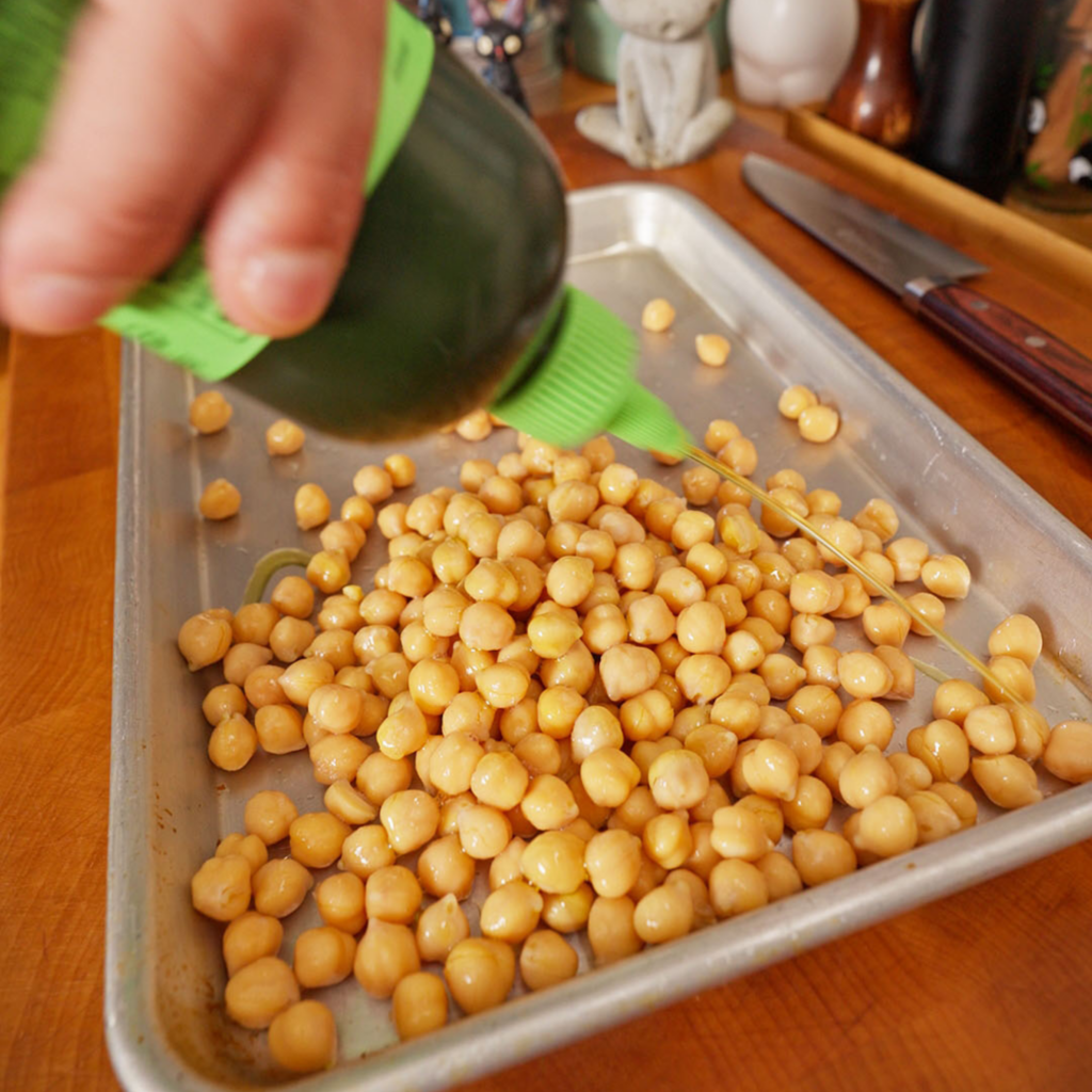 Olive oil being drizzled from a green squeeze bottle over chickpeas on a sheet pan, coating the beans evenly.