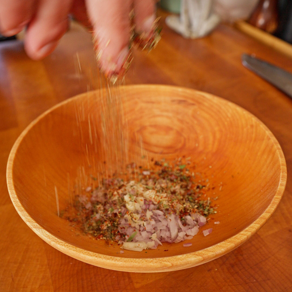 A hand sprinkling dried herbs and spices over diced red onion and garlic in a wooden bowl.