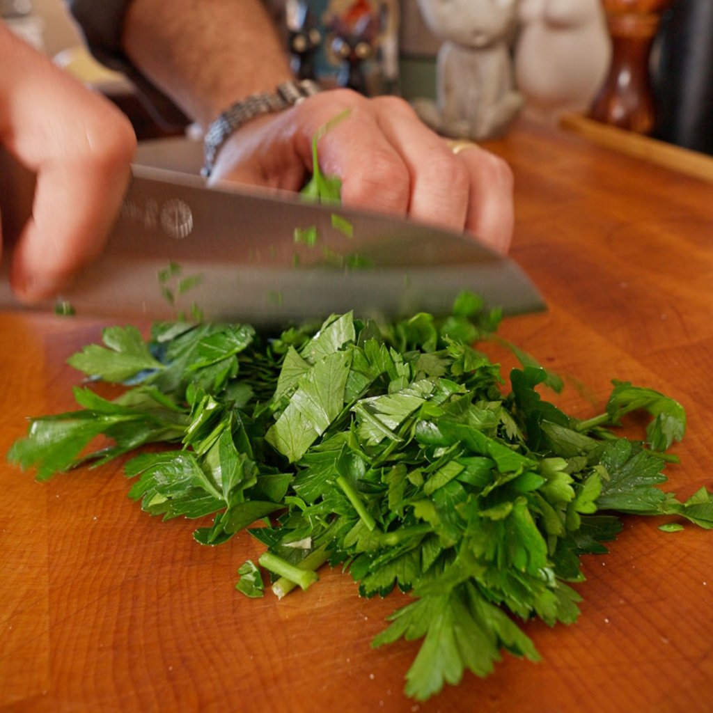 Fresh parsley being chopped with a chef’s knife on a wooden cutting board, with a pile of green leaves gathered under the blade.