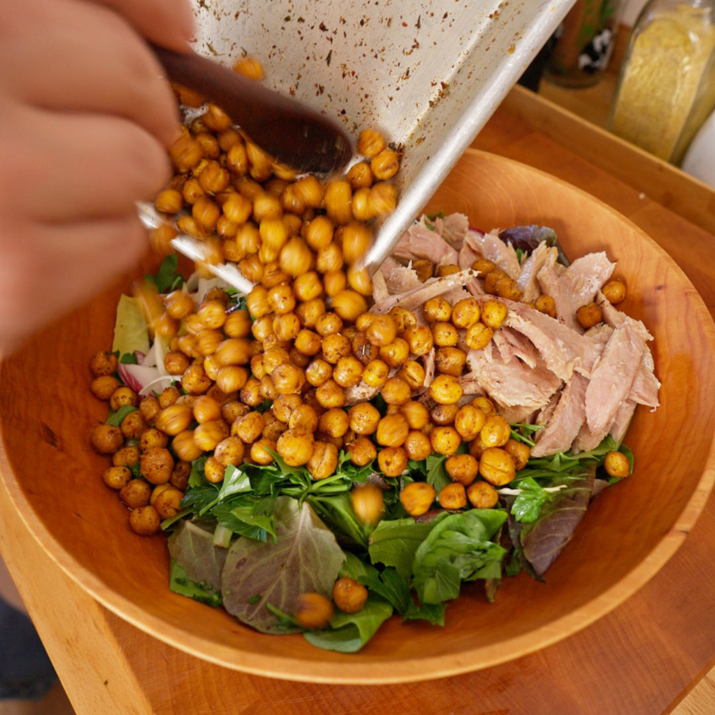 Crispy roasted chickpeas being poured from a sheet pan into a wooden bowl with mixed greens and flaked tuna.