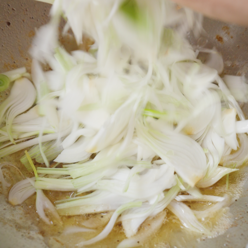 Sliced spring onions are dropped into a pan with hot rendered fat, beginning to soften and sizzle.