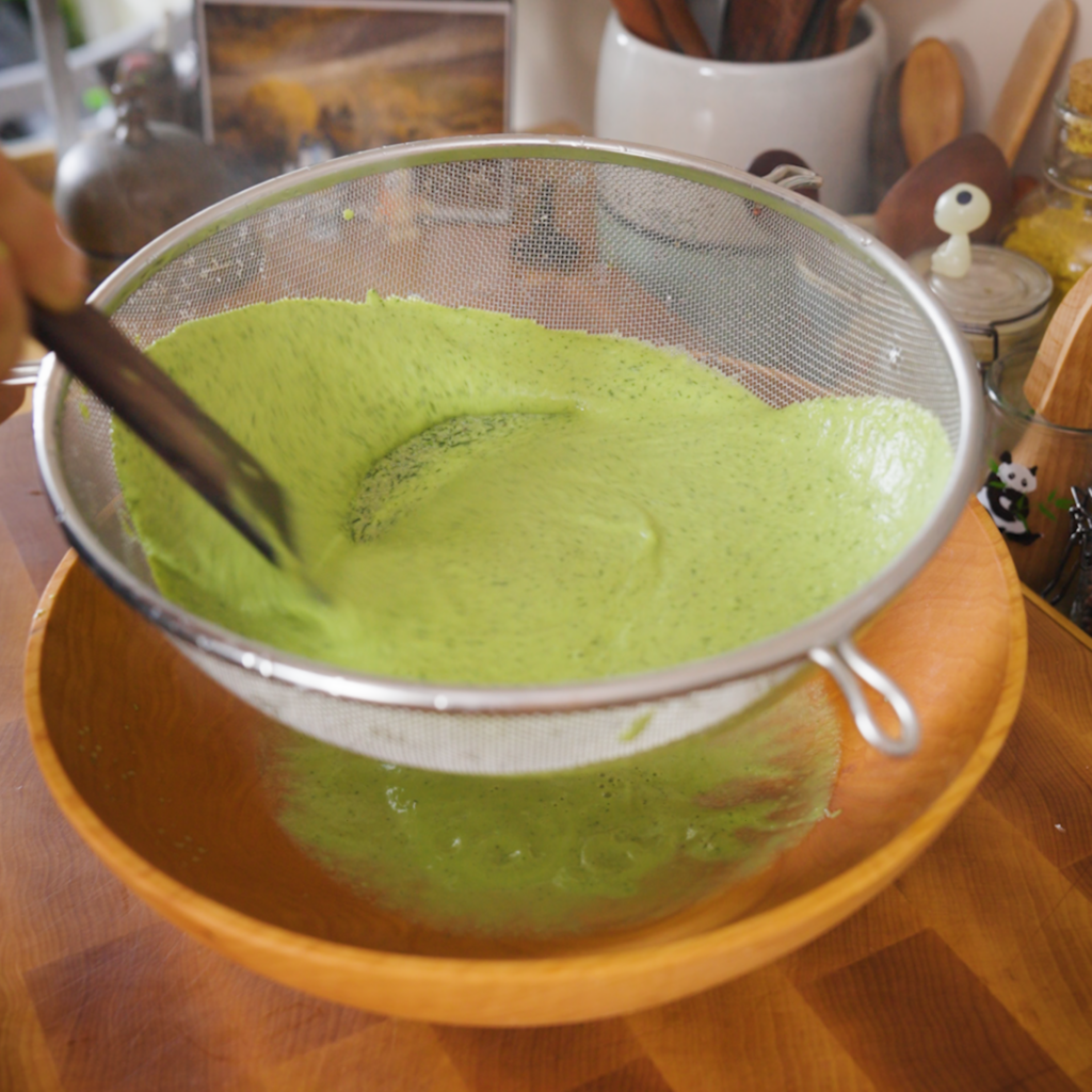 Bright green blended sauce is poured through a fine mesh strainer into a bowl, with a utensil helping push it through.