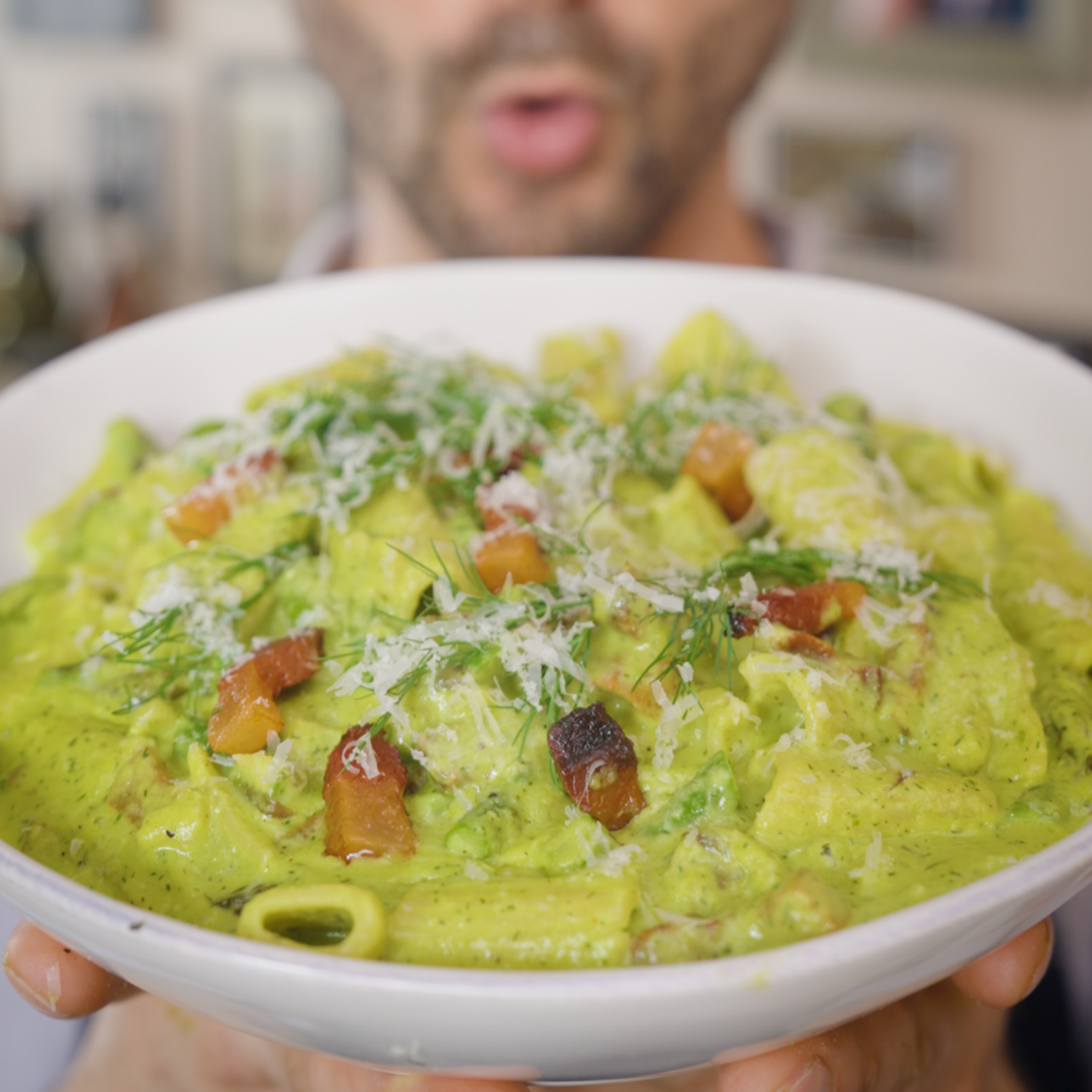 A person holds a wide bowl of rigatoni in creamy green sauce topped with crispy guanciale, grated cheese, and fresh herbs, with the face blurred in the background.