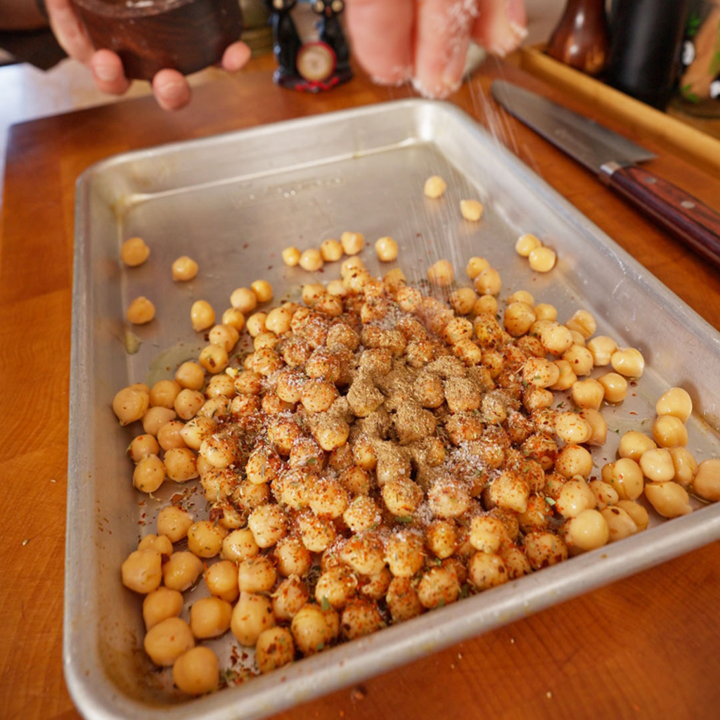Chickpeas on a sheet pan being sprinkled with salt and a mix of spices, including red chili flakes and ground seasonings, from a hand above.