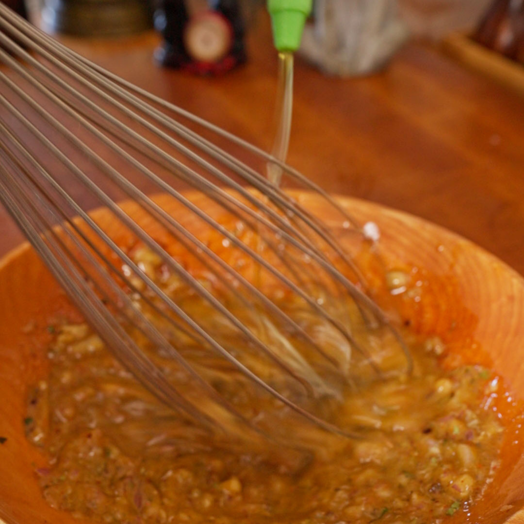 Olive oil being poured into a wooden bowl while a whisk mixes a dressing with chopped onion, garlic, and spices until combined.