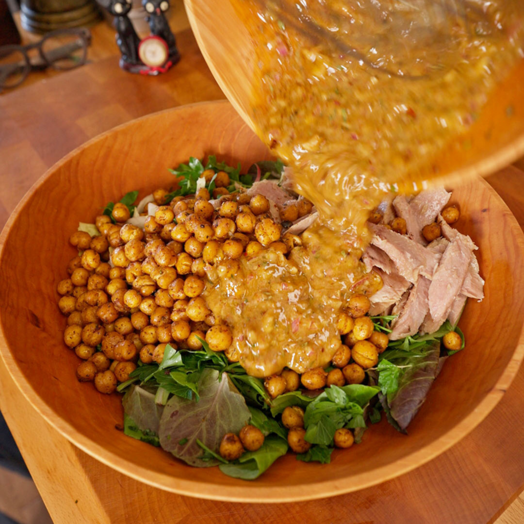 A chunky golden dressing with visible shallots and herbs being poured over a salad with chickpeas, tuna, and greens in a wooden bowl.