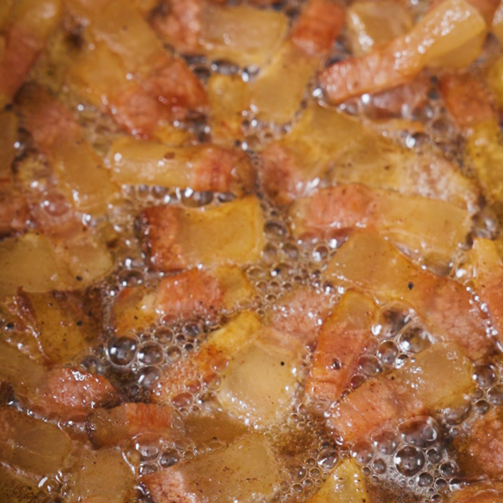 Cubed guanciale sizzling in a pan, releasing fat as the pieces turn golden brown and bubble in rendered oil.