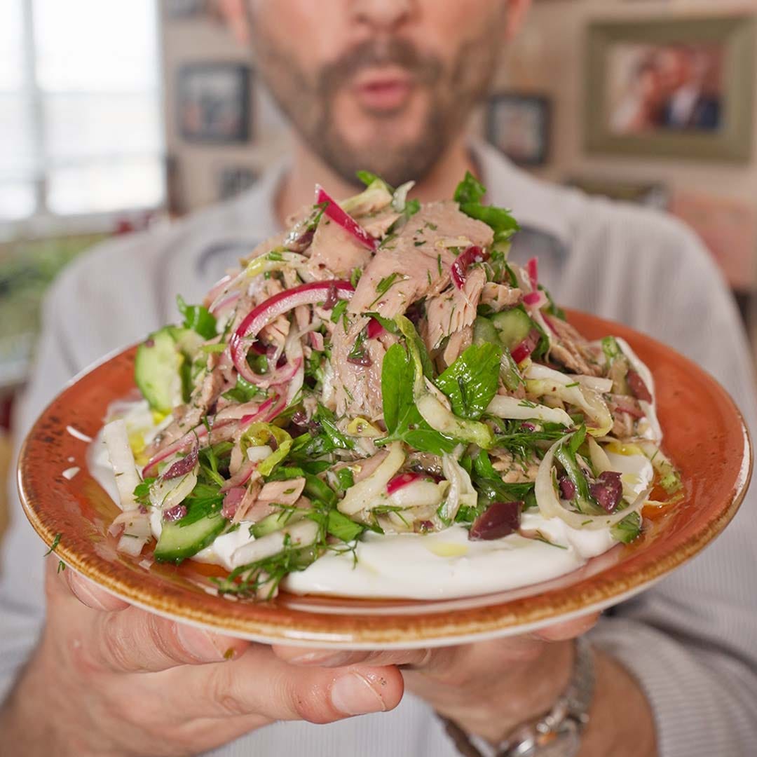 A person holds a brown plate piled with tuna salad over a thick layer of white yogurt or labneh, with sliced red onion, cucumber, herbs, and dark olives visible.