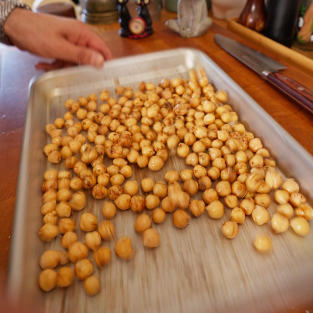 Sheet pan of chickpeas being shaken to distribute oil and spices evenly, with the chickpeas moving across the surface of the pan.