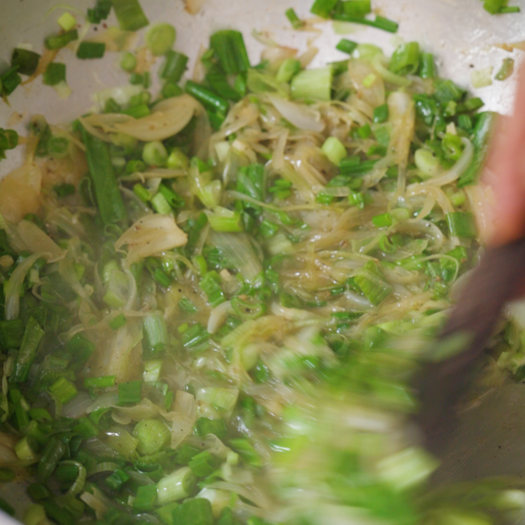 Spring onions cook down in a pan, turning soft and glossy as they are stirred with a utensil.