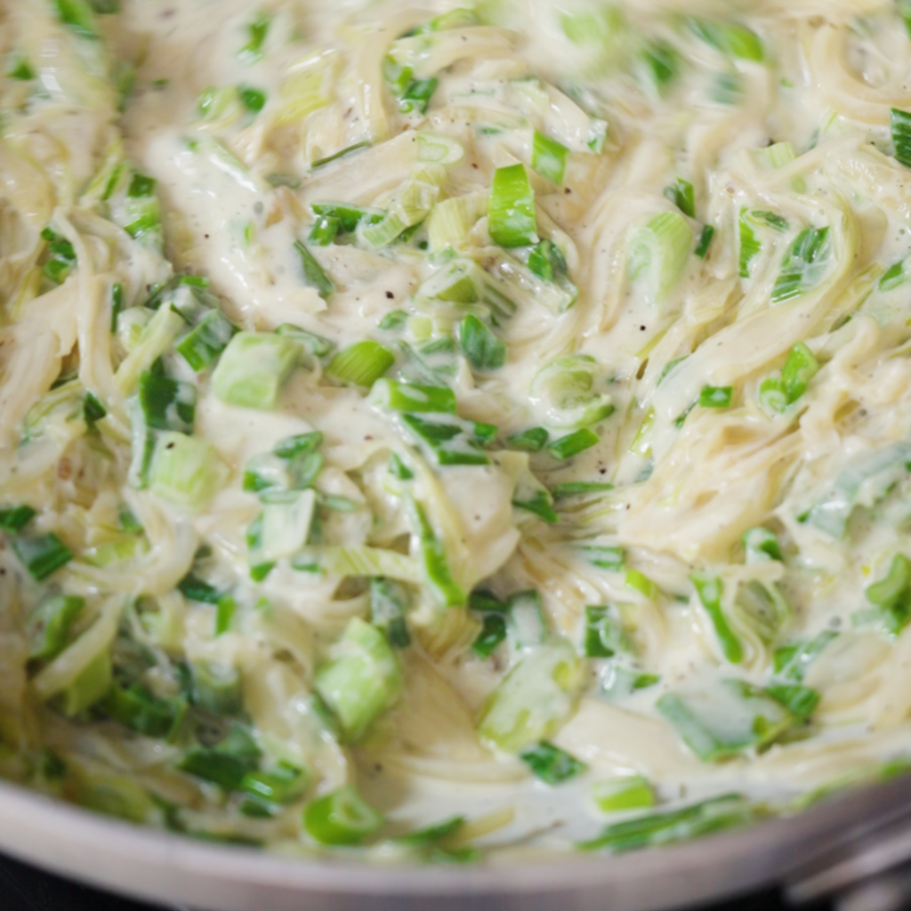 Close-up of creamy pasta sauce with cream and softened spring onions and green flecks, thick and glossy in a pan.