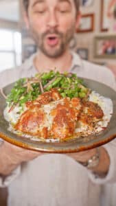 A person holds a dark ceramic plate topped with browned herb-marinated chicken thighs over a thick layer of white sauce, with a green snap pea salad and sliced red onions on the side.