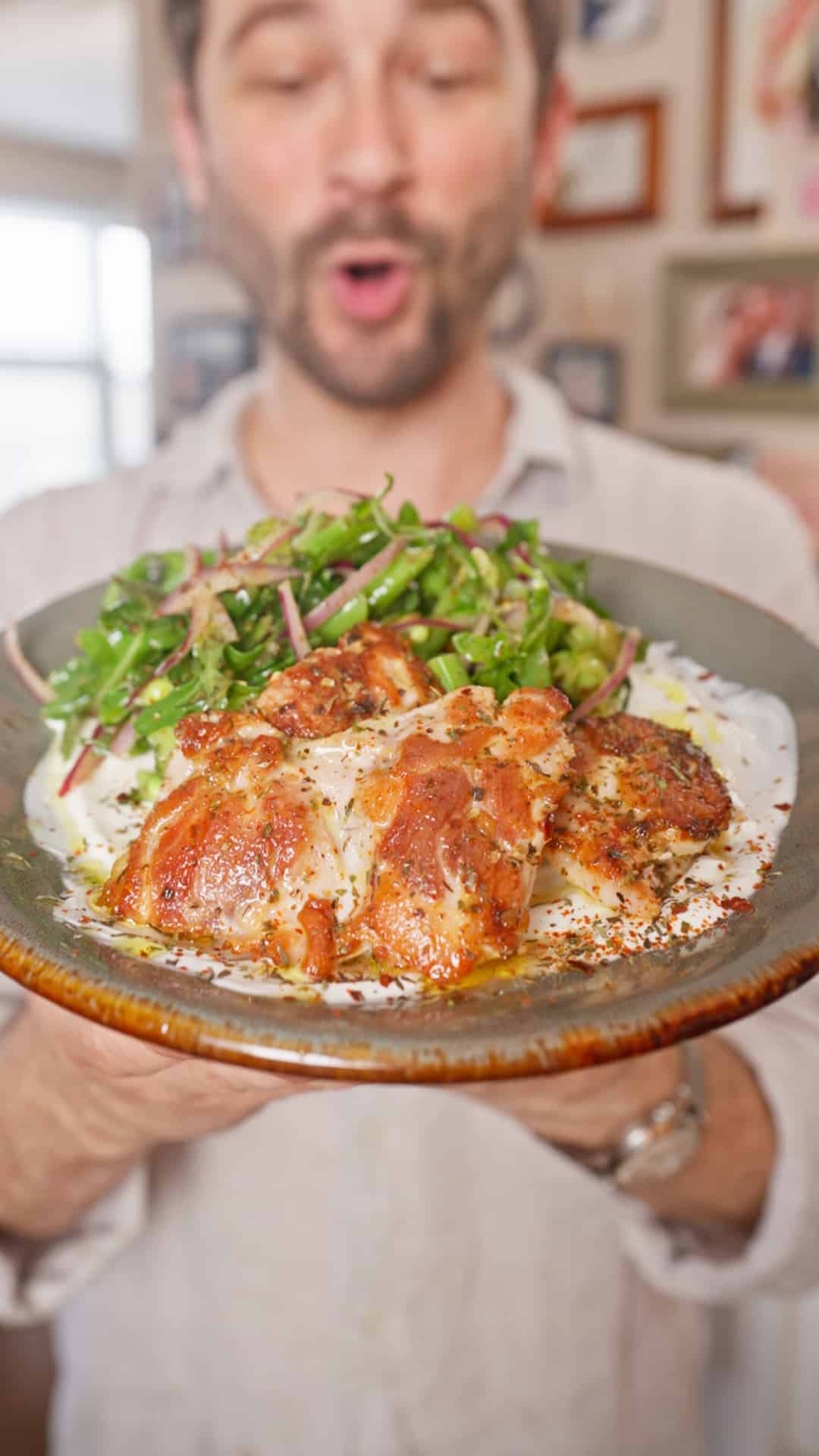 A person holds a dark ceramic plate topped with browned herb-marinated chicken thighs over a thick layer of white sauce, with a green snap pea salad and sliced red onions on the side.