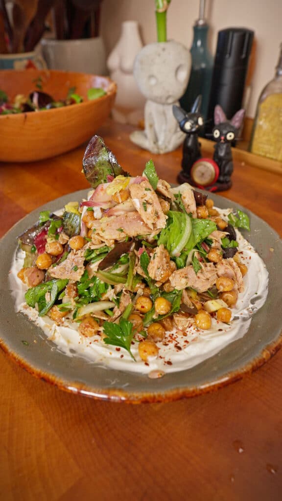 A ceramic plate filled with a yogurt base topped with flaked tuna, crispy chickpeas, leafy greens, sliced red onion, and fresh herbs, set on a wooden table with a salad bowl and small kitchen decor in the background.