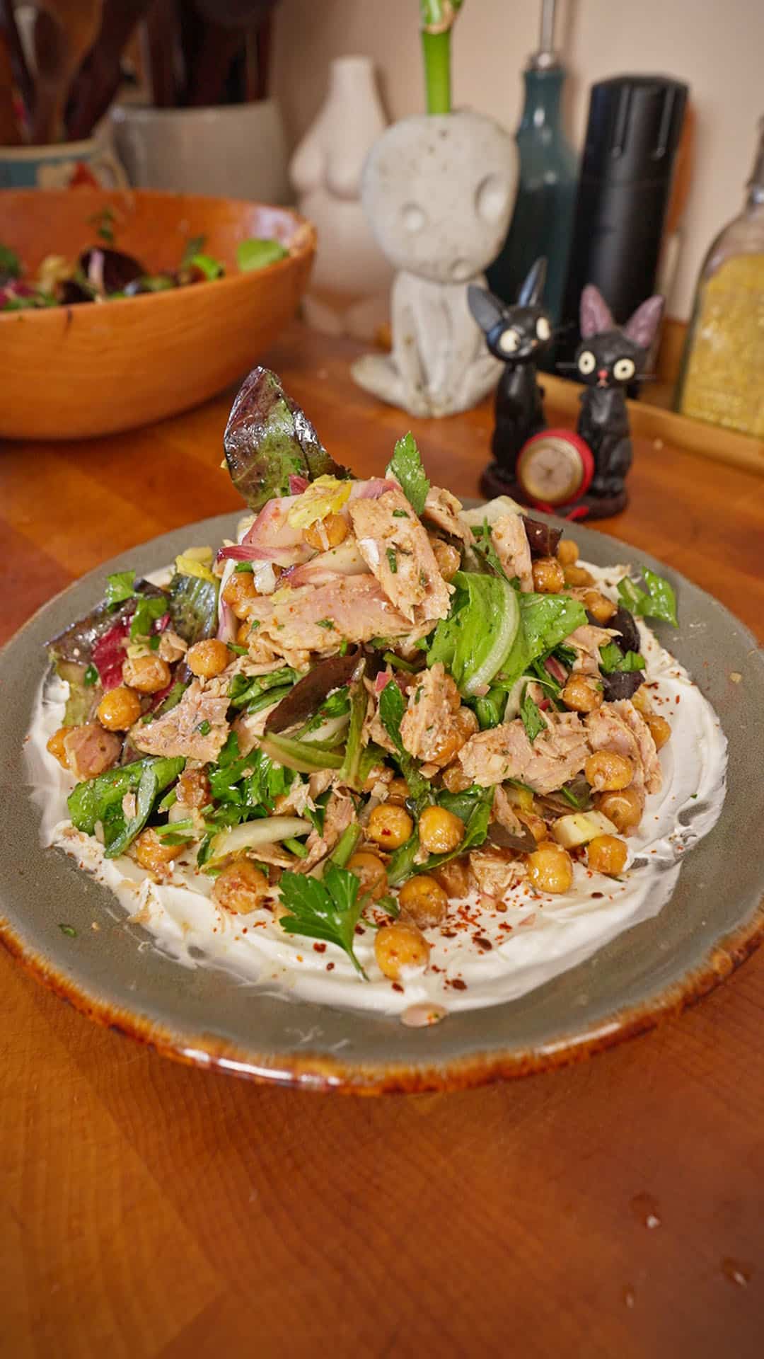 A ceramic plate filled with a yogurt base topped with flaked tuna, crispy chickpeas, leafy greens, sliced red onion, and fresh herbs, set on a wooden table with a salad bowl and small kitchen decor in the background.