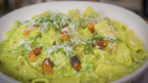 A wide white bowl filled with rigatoni coated in a creamy green sauce, topped with crispy guanciale pieces, grated cheese, and fresh dill.