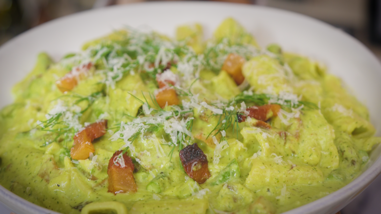 A wide white bowl filled with rigatoni coated in a creamy green sauce, topped with crispy guanciale pieces, grated cheese, and fresh dill.