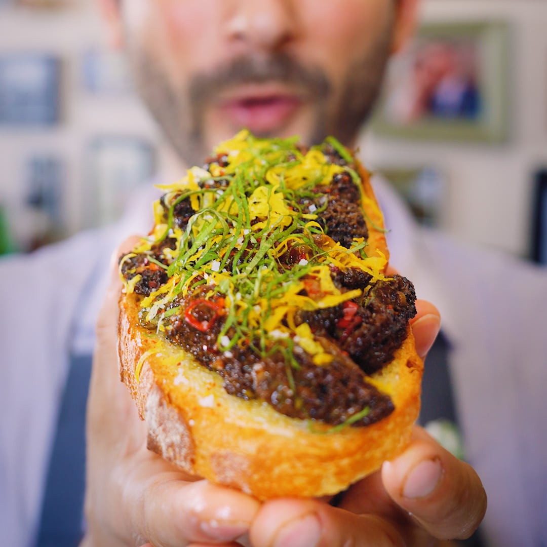A hand holds a slice of toasted bread topped with sautéed morel mushrooms, thinly sliced herbs, and lemon zest, with a blurred face in the background.