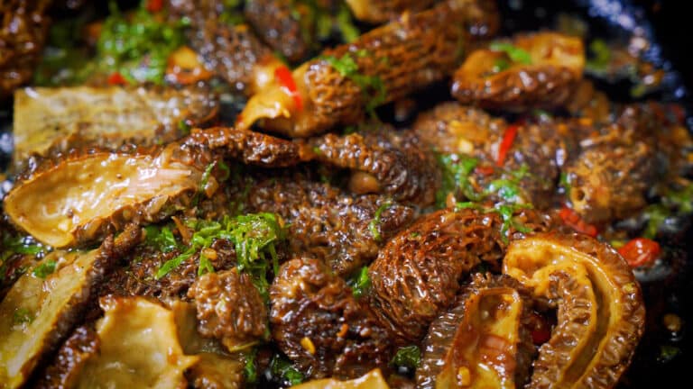 Close-up of morel mushrooms cooking in a pan, showing their textured caps coated in a rich, buttery sauce with herbs.